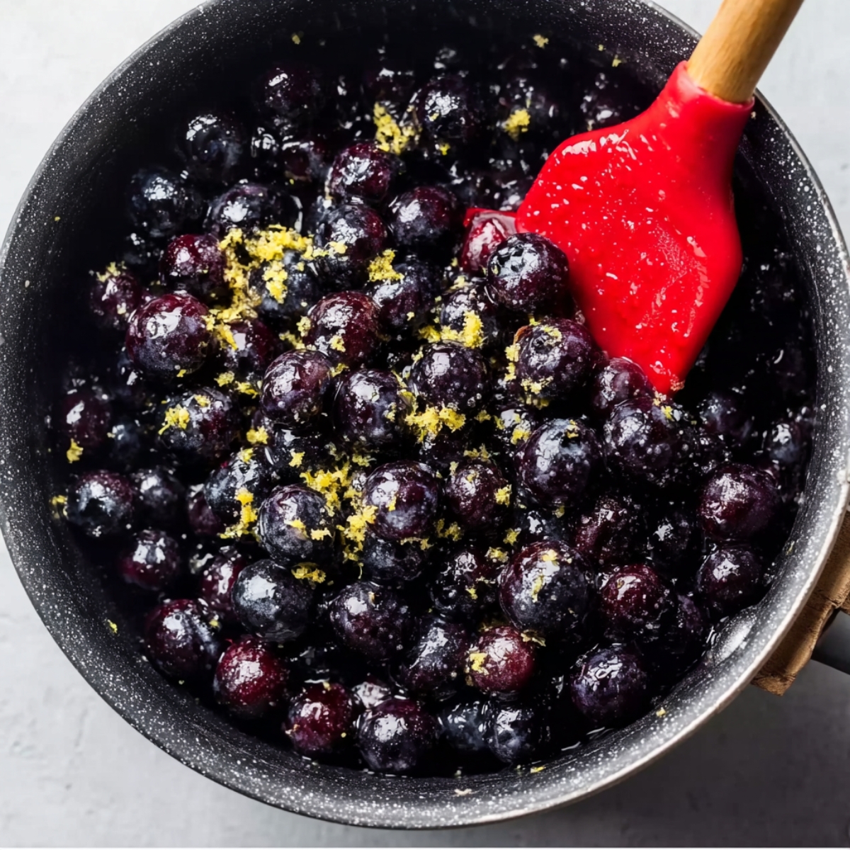 Easy Blueberry Pie Bars Recipe 11 Fresh blueberries cooking in a saucepan with lemon zest and a red silicone spatula, close-up homemade blueberry filling preparation on a marble surface.