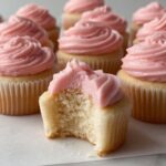 Copycat Magnolia Bakery Cupcakes with swirled pink frosting arranged on a white surface, one cupcake in the foreground with a bite taken to reveal the soft interior.