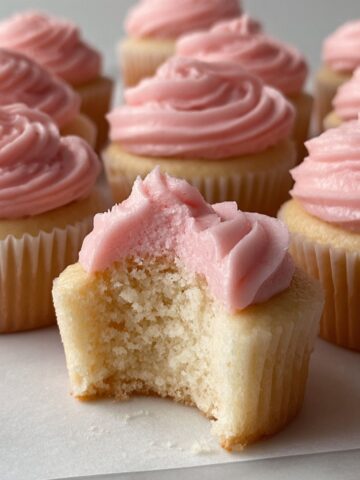 Copycat Magnolia Bakery Cupcakes 1 Copycat Magnolia Bakery Cupcakes with swirled pink frosting arranged on a white surface, one cupcake in the foreground with a bite taken to reveal the soft interior.