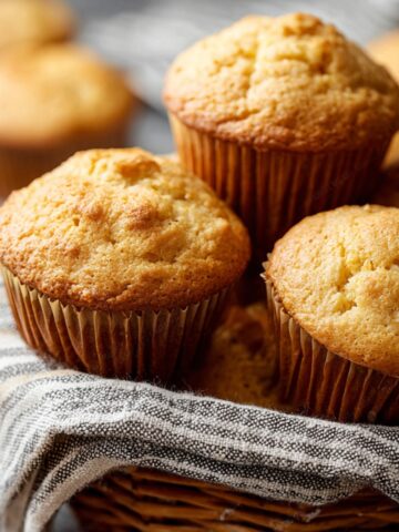 The Best Cornbread Muffins Recipe 1 Golden-brown baked cornbread muffins in paper liners, fresh out of the oven and arranged in a basket lined with a striped cloth.
