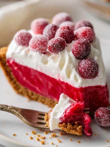 Best Cranberry Curd Tart Recipe 1 Slice of Cranberry Curd Tart with graham cracker crust, topped with whipped cream and sugared cranberries on a white plate with a fork.