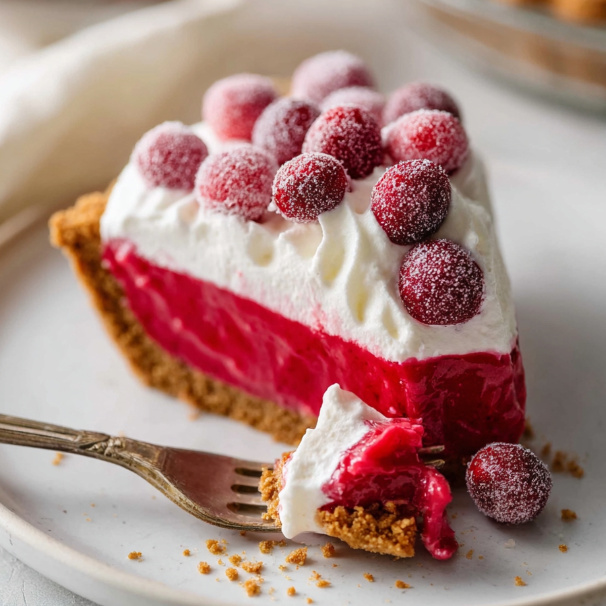 Slice of Cranberry Curd Tart with graham cracker crust, topped with whipped cream and sugared cranberries on a white plate with a fork.