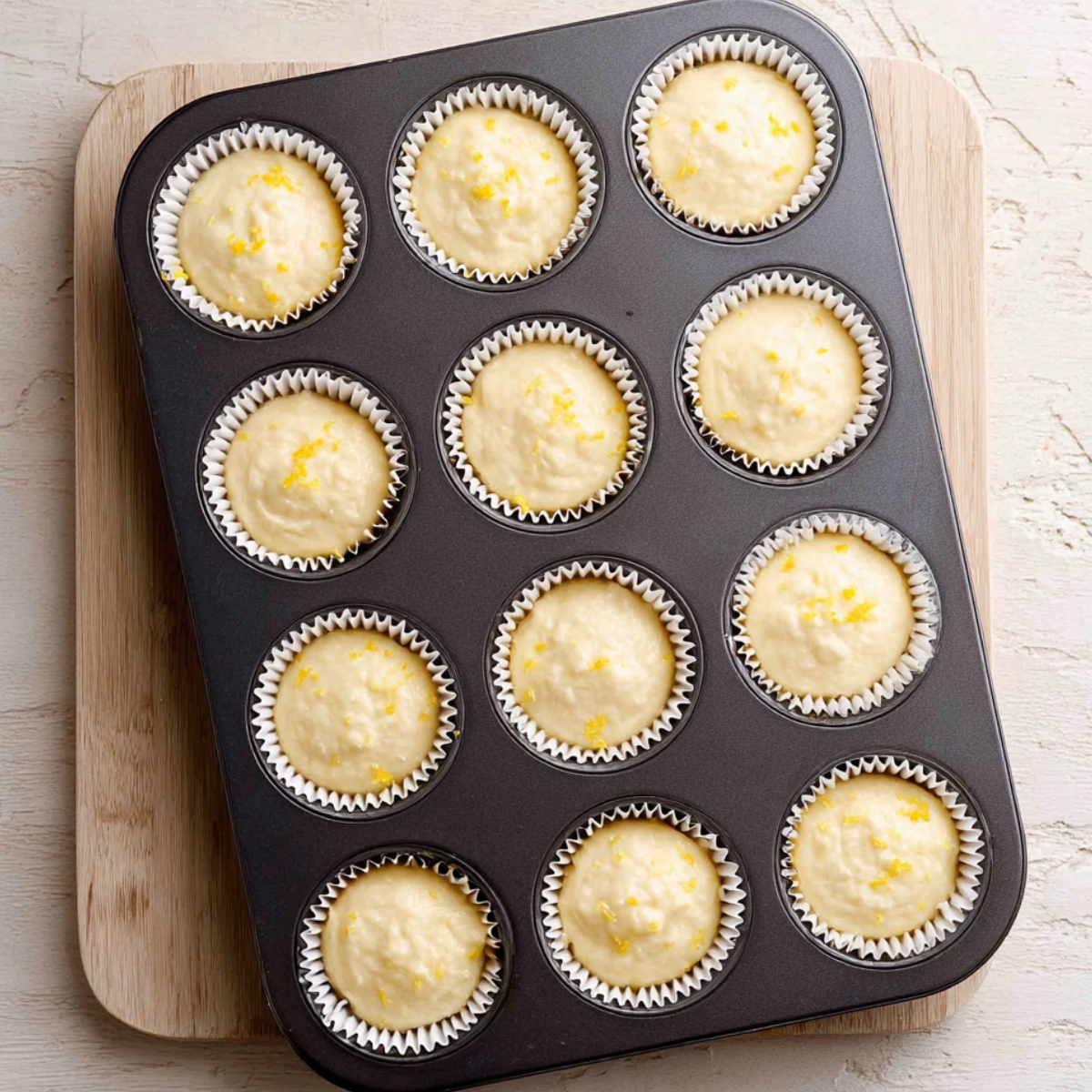 Best Lemon Raspberry Jam Cupcakes 10 A 12-cup muffin tin filled with lemon cupcake batter in white paper liners, sprinkled with lemon zest, ready to bake.