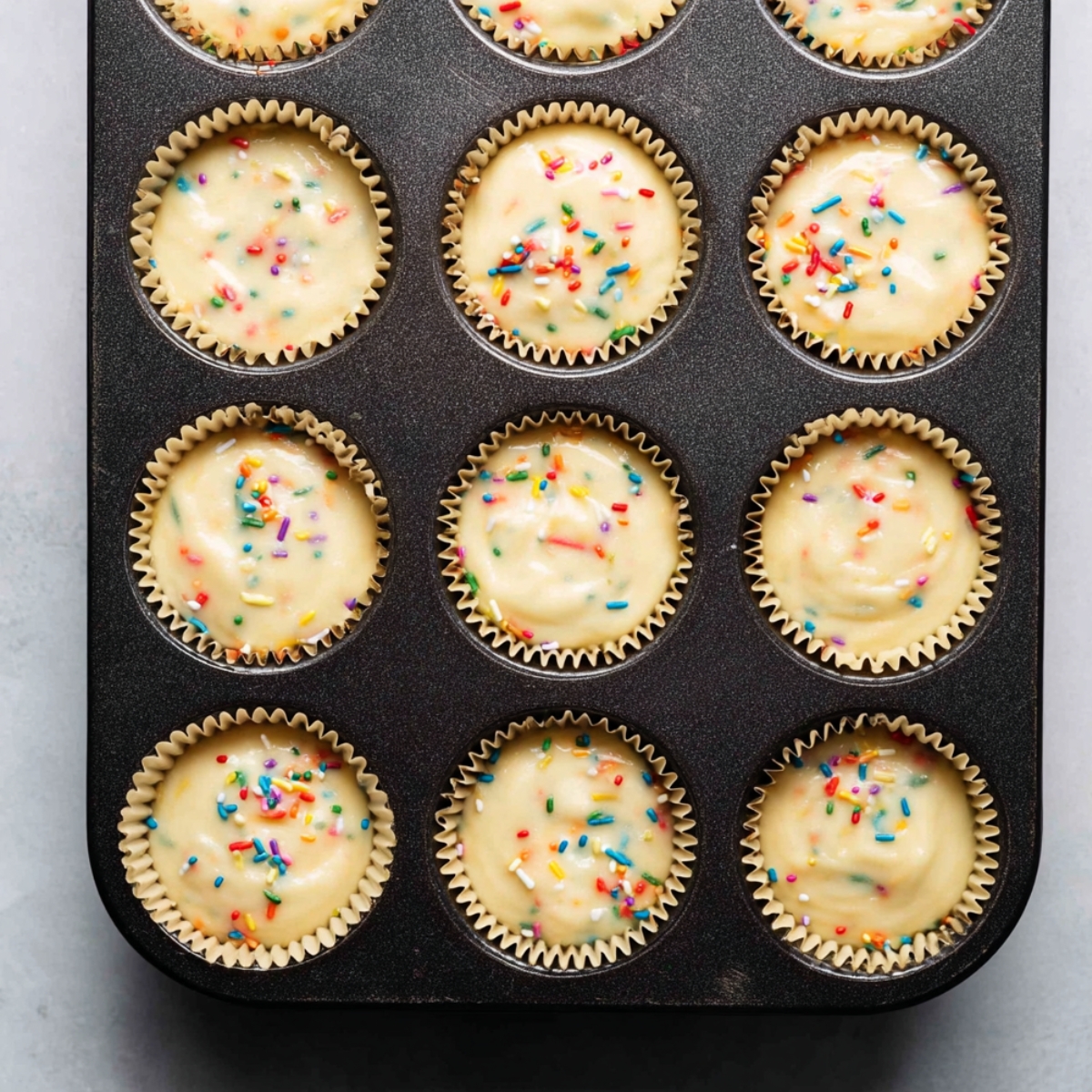 Easy Confetti Sprinkle Cupcakes Recipe 11 Funfetti cupcake batter with colorful rainbow sprinkles portioned into paper cupcake liners inside a dark muffin tin.