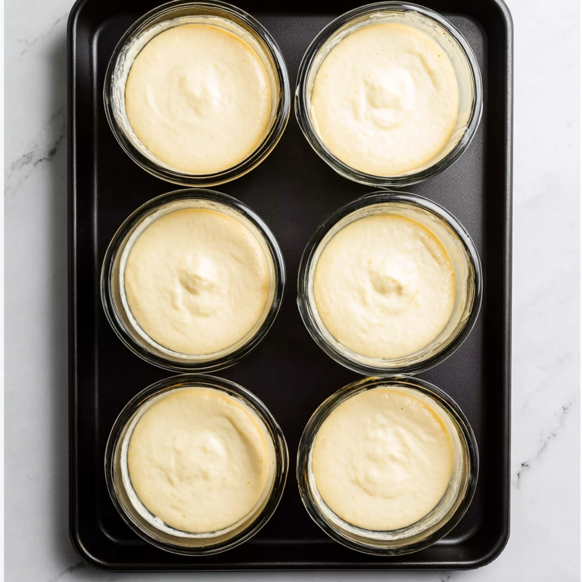 Easy Lemon Pudding Cakes Recipe 11 Lemon pudding cake batter in glass ramekins on a black tray.
