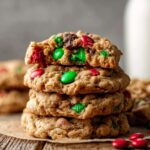 Stack of baked Flourless Monster Cookies with red and green M&M’s and chocolate chips, one cookie on top is broken in half showing the chewy interior.