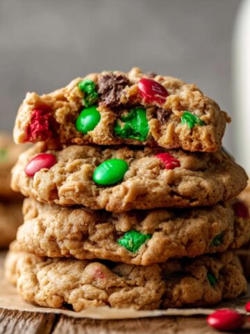 Stack of baked Flourless Monster Cookies with red and green M&M’s and chocolate chips, one cookie on top is broken in half showing the chewy interior.