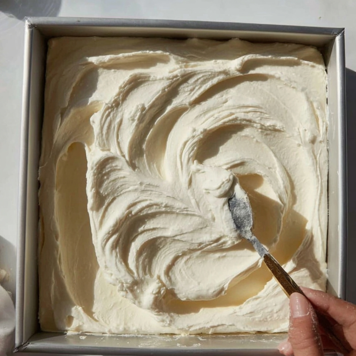 Easy Coconut Cream Bars Recipe 11 Close-up of a square dessert pan being frosted with creamy white icing using a spoon, showing smooth, swirled texture on top of the dessert.