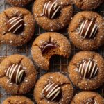 Freshly baked Gingerbread Blossoms cookies on a cooling rack, topped with chocolate kisses and drizzled white chocolate, with one cookie showing a bite to reveal the soft interior.