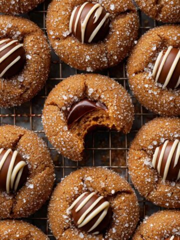 The Best Gingerbread Blossoms Recipe 2 Freshly baked Gingerbread Blossoms cookies on a cooling rack, topped with chocolate kisses and drizzled white chocolate, with one cookie showing a bite to reveal the soft interior.