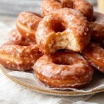 Stack of freshly made Homemade Glazed Doughnuts on a plate, one with a bite taken out showing the soft, fluffy interior, with shiny sugar glaze coating.