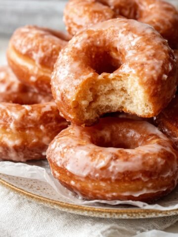 Homemade Glazed Doughnuts Recipe 3 Stack of freshly made Homemade Glazed Doughnuts on a plate, one with a bite taken out showing the soft, fluffy interior, with shiny sugar glaze coating.