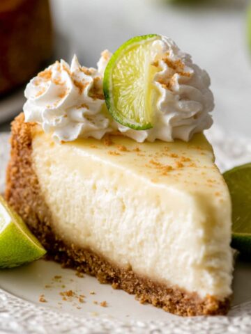 The Best Key Lime Cheesecake Recipe 2 Slice of creamy Key Lime Cheesecake on a white plate, topped with whipped cream and fresh lime wedges, with a slice of lime cheesecake in the background.