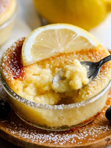 Easy Lemon Pudding Cakes Recipe 2 Baked Lemon Pudding Cakes in a ramekin with lemon slice and blueberries, spoon scooping.