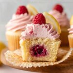 Lemon Raspberry Jam Cupcakes with pink raspberry frosting, topped with fresh raspberries and lemon wedges. One cupcake is cut open to show a juicy raspberry jam filling inside.