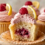 Lemon Raspberry Jam Cupcakes with pink raspberry frosting, topped with fresh raspberries and lemon wedges. One cupcake is cut open to show a juicy raspberry jam filling inside.