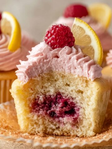 Best Lemon Raspberry Jam Cupcakes 4 Lemon Raspberry Jam Cupcakes with pink raspberry frosting, topped with fresh raspberries and lemon wedges. One cupcake is cut open to show a juicy raspberry jam filling inside.