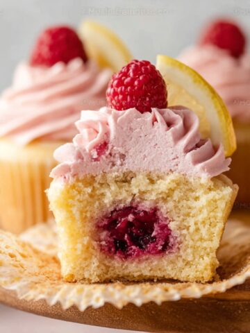 Best Lemon Raspberry Jam Cupcakes 1 Lemon Raspberry Jam Cupcakes with pink raspberry frosting, topped with fresh raspberries and lemon wedges. One cupcake is cut open to show a juicy raspberry jam filling inside.