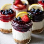 No Bake Cheesecake Jars layered with graham cracker crust, creamy filling, and berry topping garnished with strawberries, blueberries, and lemon slices.
