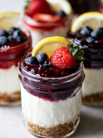 Easy No Bake Cheesecake Jars Recipe 1 No Bake Cheesecake Jars layered with graham cracker crust, creamy filling, and berry topping garnished with strawberries, blueberries, and lemon slices.