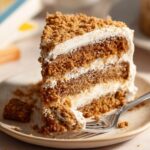 A slice of Oatmeal Cream Pie Cake on a plate, layered with creamy frosting and topped with crumbled oatmeal cookies, with a fork taking a bite.