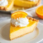 Slice of creamy Orange Pie topped with whipped cream and a fresh orange slice on a white plate with whole pie blurred in the background.