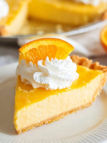 The Best Orange Pie Recipe 1 Slice of creamy Orange Pie topped with whipped cream and a fresh orange slice on a white plate with whole pie blurred in the background.