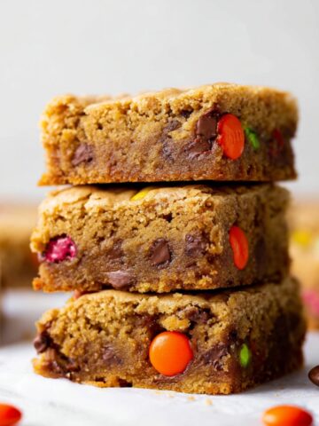 A stack of three baked Peanut Butter Blondies with chocolate chips and candy pieces, surrounded by scattered candies on parchment paper.