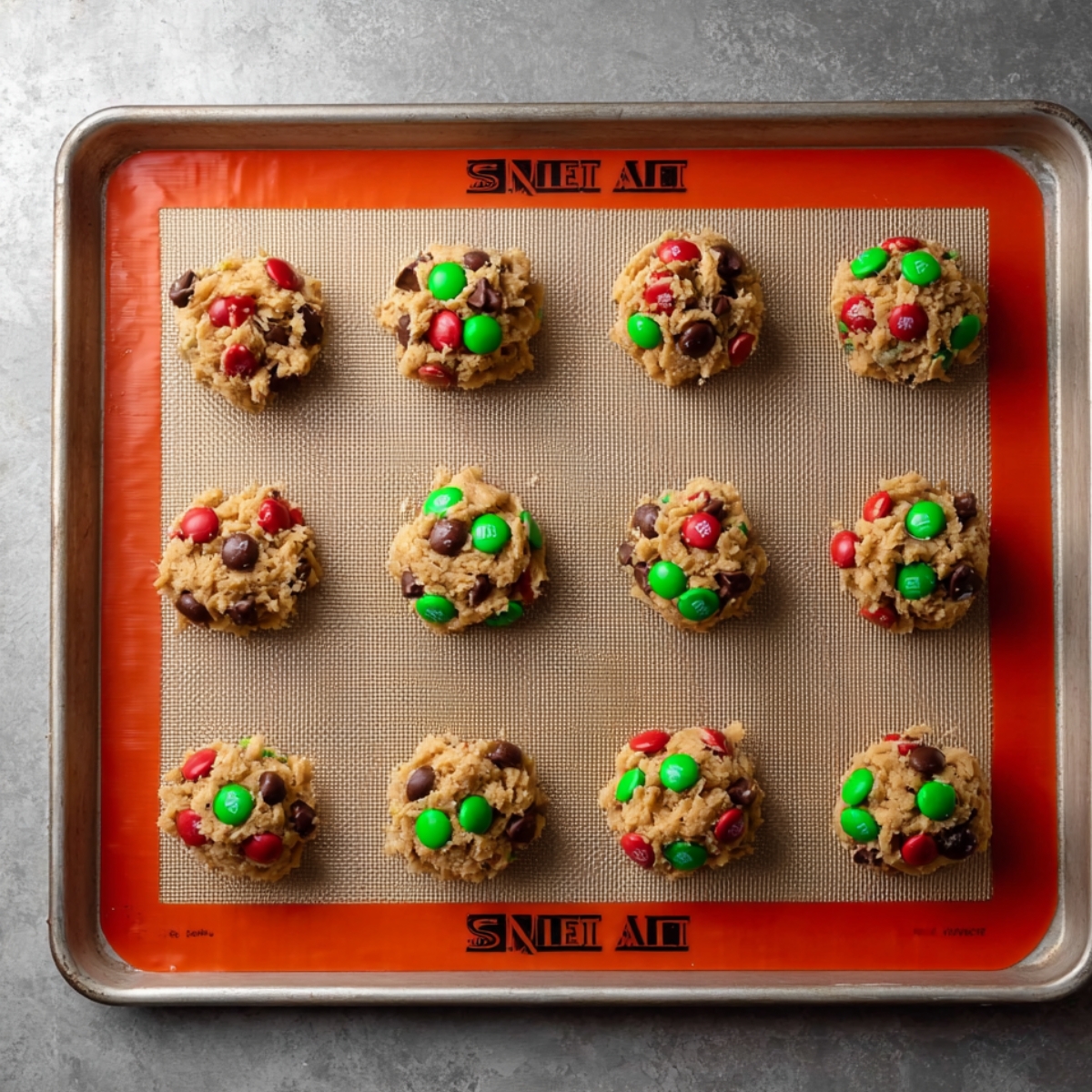 Easy Flourless Monster Cookies Recipe 11 Close-up of cookie dough scoops with red and green M&M’s and chocolate chips on a baking sheet ready to bake.