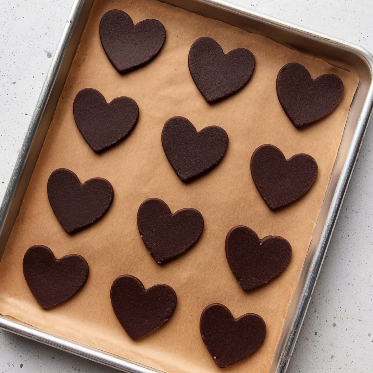 Cut-Out Chocolate Sugar Cookies 11 A tray lined with parchment paper containing freshly cut heart-shaped chocolate sugar cookies, ready to be baked. The cookies are neatly arranged in rows, showing a smooth surface.