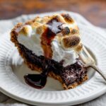 Slice of S'mores Brownie Pie topped with toasted marshmallows and chocolate sauce on a white dessert plate.