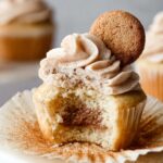 A close-up of a Snickerdoodle Cupcakes with a bite taken out, revealing the cinnamon swirl inside. The cupcake is topped with a swirl of cinnamon buttercream frosting and a cinnamon sugar cookie.