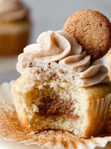 Snickerdoodle Cupcakes with Cinnamon Swirl Frosting 1 A close-up of a Snickerdoodle Cupcakes with a bite taken out, revealing the cinnamon swirl inside. The cupcake is topped with a swirl of cinnamon buttercream frosting and a cinnamon sugar cookie.