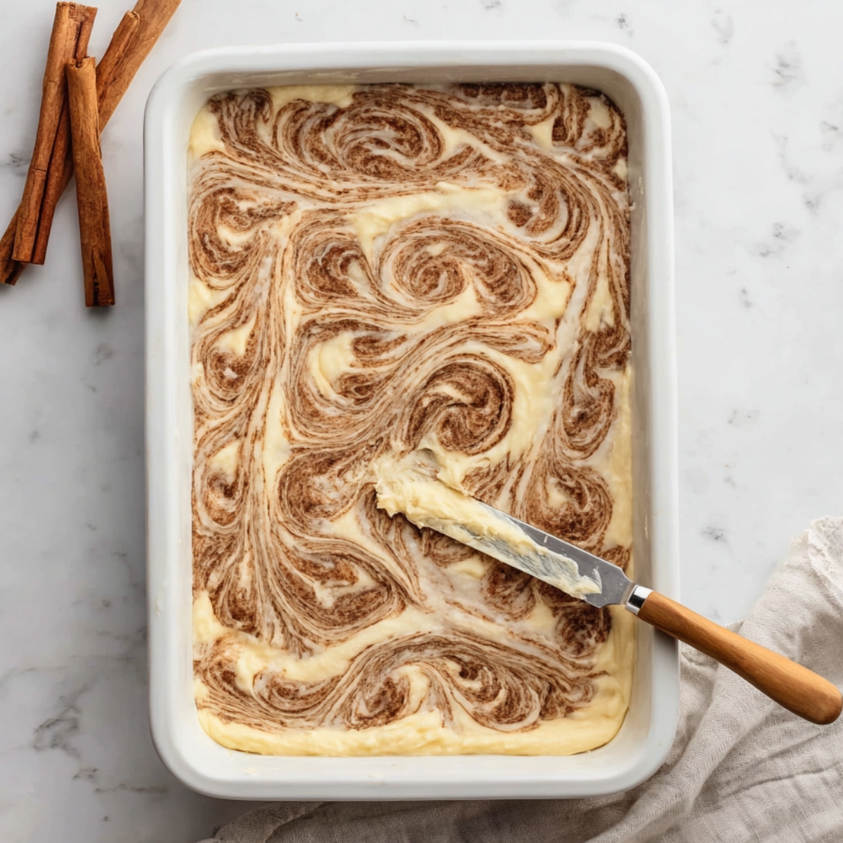 Cinnamon Roll Coffee Cake Recipe 11 Overhead view of cinnamon sugar filling swirled through cake batter in a white baking dish, creating a marbled cinnamon roll pattern.
