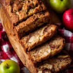Thick slices of homemade Apple Cinnamon Quick Bread with tender apple chunks and cinnamon swirl, served on a wooden board with fresh apples and a red checkered cloth.