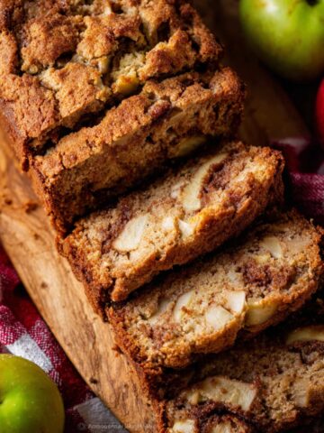 Apple Cinnamon Quick Bread Recipe 1 Thick slices of homemade Apple Cinnamon Quick Bread with tender apple chunks and cinnamon swirl, served on a wooden board with fresh apples and a red checkered cloth.