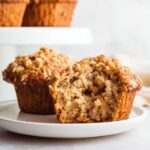 Moist Apple Oatmeal Muffins cut in half on a white plate, showing tender crumb, apple pieces, and crunchy oat streusel topping, with more muffins blurred in the background.