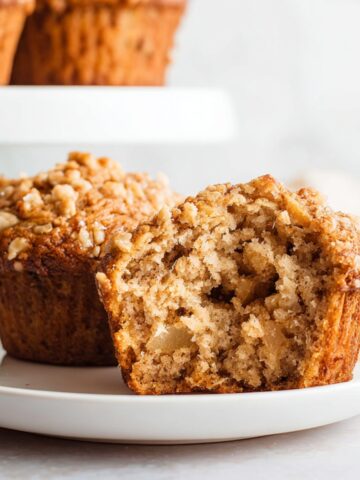 Easy Apple Oatmeal Muffins Recipe 1 Moist Apple Oatmeal Muffins cut in half on a white plate, showing tender crumb, apple pieces, and crunchy oat streusel topping, with more muffins blurred in the background.
