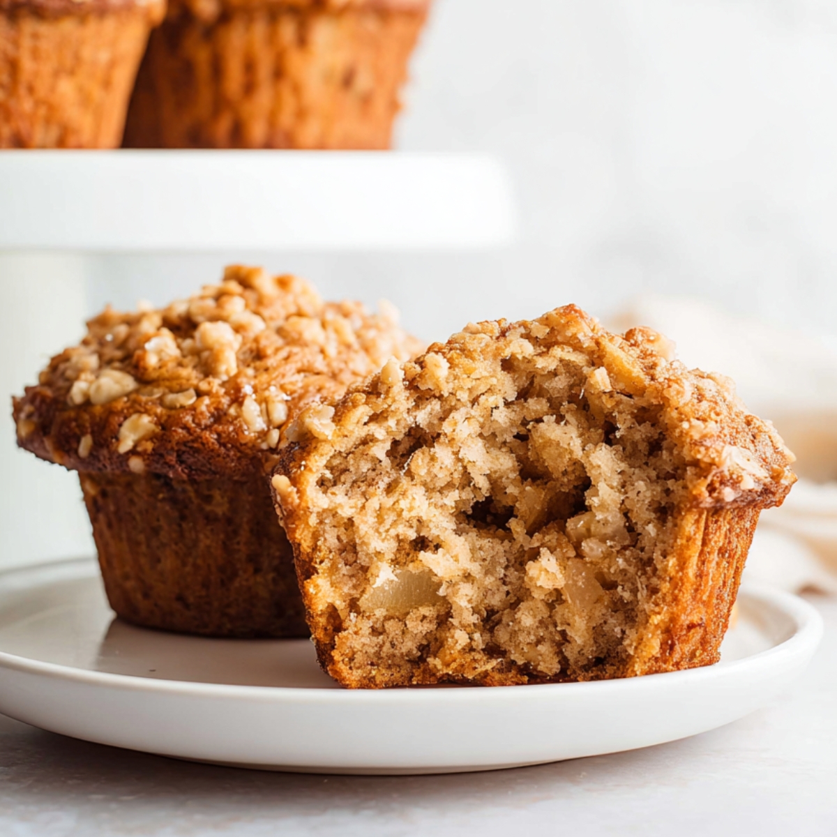 Easy Apple Oatmeal Muffins Recipe 9 Moist Apple Oatmeal Muffins cut in half on a white plate, showing tender crumb, apple pieces, and crunchy oat streusel topping, with more muffins blurred in the background.