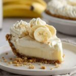 Slice of Banana Cream Cheesecake topped with whipped cream rosettes and fresh banana slices on a white plate, with whole cheesecake and bananas blurred in the background.