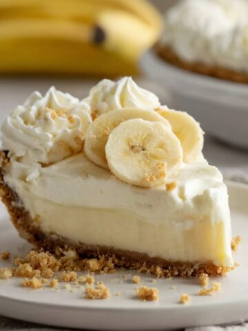 No-Bake Banana Cream Cheesecake 2 Slice of Banana Cream Cheesecake topped with whipped cream rosettes and fresh banana slices on a white plate, with whole cheesecake and bananas blurred in the background.