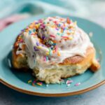 Birthday Cake Cinnamon Rolls topped with thick vanilla frosting and rainbow sprinkles on a pastel blue plate, soft fluffy pastry texture, cozy bakery-style dessert photography.