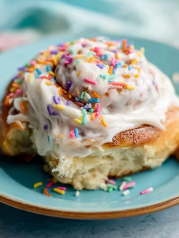 Birthday Cake Cinnamon Rolls Recipe 1 Birthday Cake Cinnamon Rolls topped with thick vanilla frosting and rainbow sprinkles on a pastel blue plate, soft fluffy pastry texture, cozy bakery-style dessert photography.
