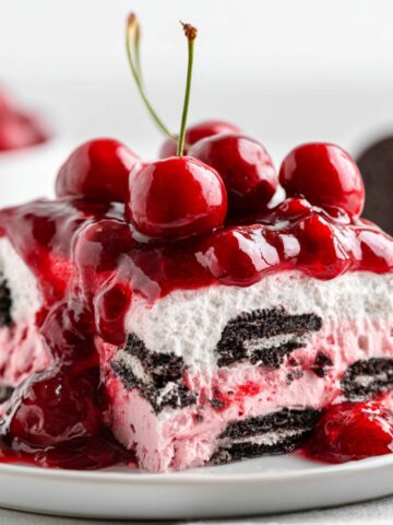 Easy Black Forest Icebox Cake Recipe 1 Slice of Black Forest Icebox Cake topped with glossy cherry pie filling and whole cherries, featuring layers of pink cream and chocolate sandwich cookies.