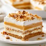 Slice of Butterscotch Lush dessert on a white plate featuring layers of graham cracker crust, cream cheese filling, butterscotch pudding, and whipped topping, finished with crushed toffee pieces.