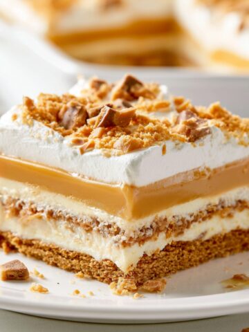 The Best Butterscotch Lush Recipe 1 Slice of Butterscotch Lush dessert on a white plate featuring layers of graham cracker crust, cream cheese filling, butterscotch pudding, and whipped topping, finished with crushed toffee pieces.