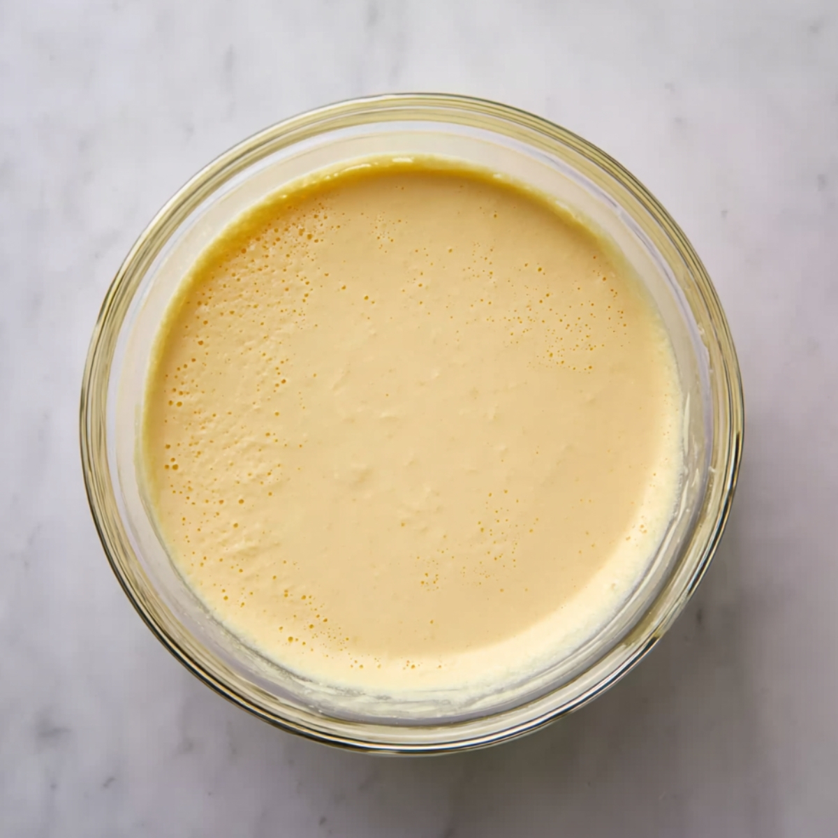 The Best Waffles Recipe 11 Smooth waffle batter in a clear glass bowl on a marble countertop, top view baking preparation scene with creamy homemade batter texture, minimalist kitchen aesthetic.