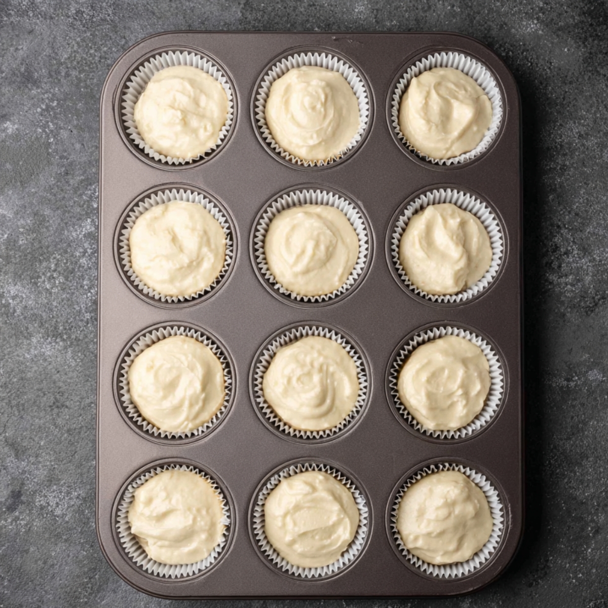 Easy Salted Caramel Cupcakes Recipe 11 Vanilla cupcake batter portioned into white paper liners in a 12-cup muffin pan, arranged on a dark stone countertop and ready for baking.