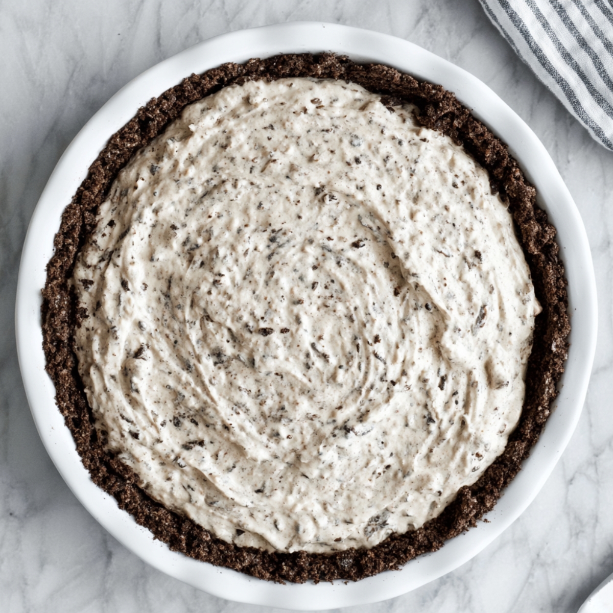 Easy Oreo Pudding Pie Recipe 11 Top-down view of a homemade Oreo pie with creamy cookies-and-cream filling spread inside a dark chocolate cookie crust in a white pie dish on a marble countertop.