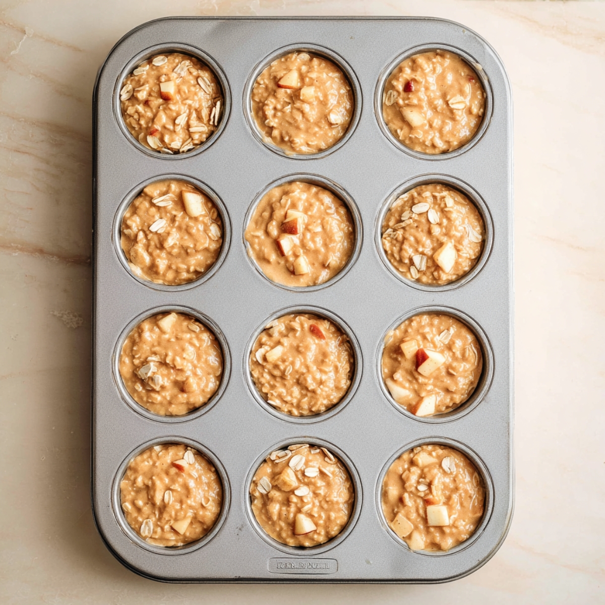 Easy Apple Oatmeal Muffins Recipe 10 Apple oatmeal muffin batter portioned into a 12-cup muffin tin, with visible apple chunks and oats, ready for baking in a home kitchen.